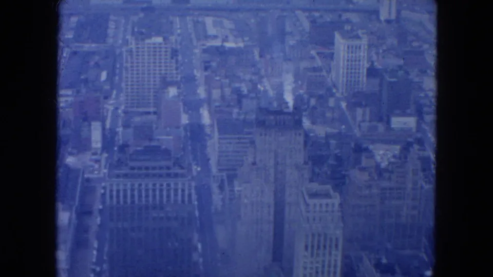 1954 New York City aerial frame showing tall commercial buildings and a traffic-lined avenue in soft blue 8mm color.