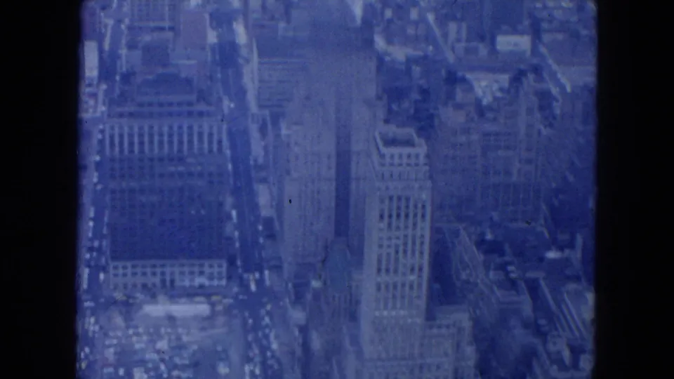 1954 New York City aerial view with office buildings, rooftops, and rows of traffic visible through blue 8mm film texture.