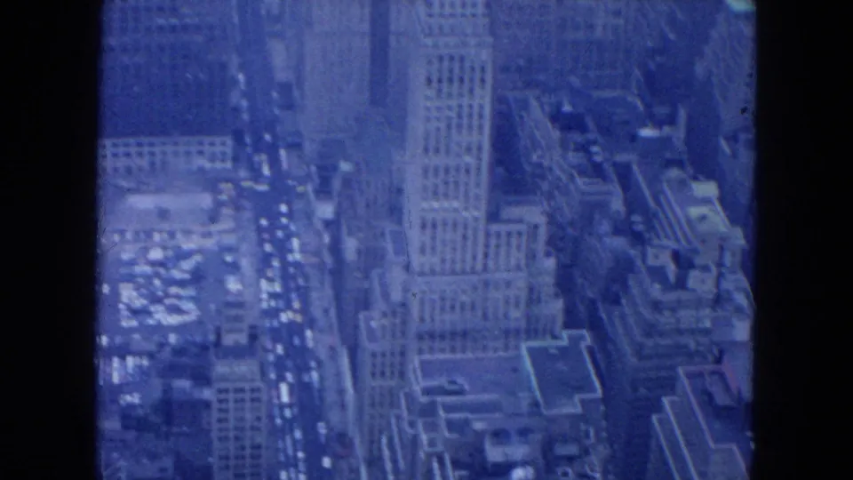 1954 New York City aerial frame looking down on a tall commercial building, nearby rooftops, and traffic moving through Midtown streets.