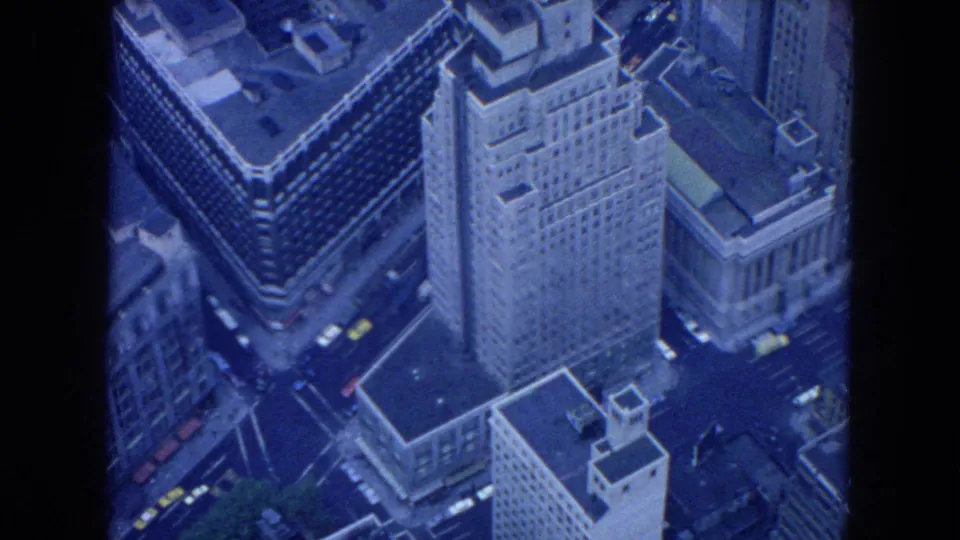 1954 New York City aerial frame showing a tall masonry building, nearby intersections, rooftops, and small vintage cars below.
