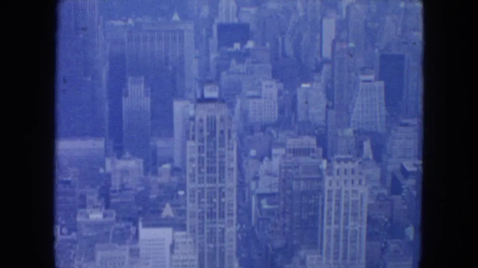 Blue-tinted 1954 New York City aerial frame showing a dense cluster of high-rise buildings and rooftops through soft 8mm exposure.
