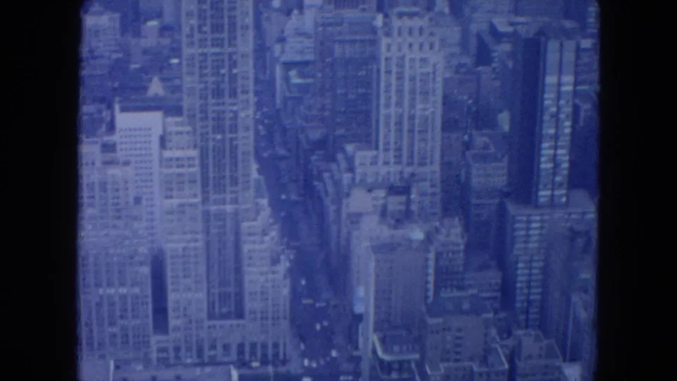 1954 New York City aerial frame looking along a street canyon between tall buildings, with small vehicles visible below.