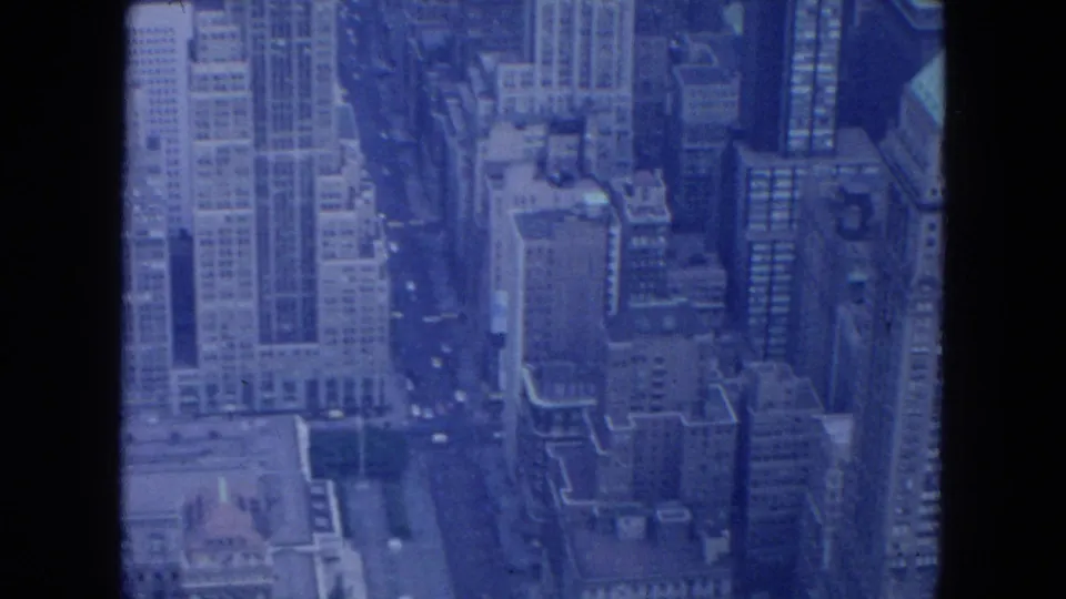 Blue-tinted 1954 New York City aerial frame showing tall office buildings, rooftops, and narrow streets below.
