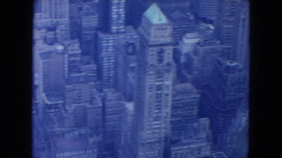 1954 New York City aerial frame with clustered towers, roof shapes, and soft blue 8mm film texture.