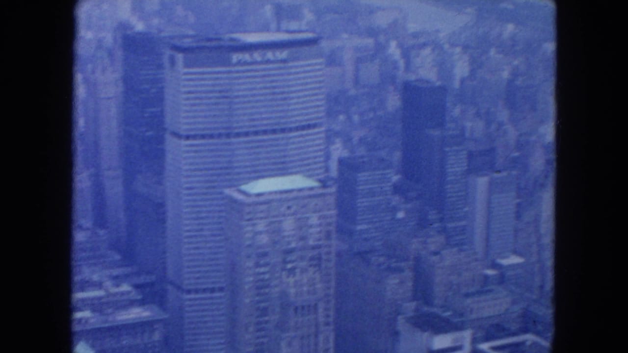 Blue-tinted aerial view of Manhattan towers with a Pan Am marking near the top of a tall building.