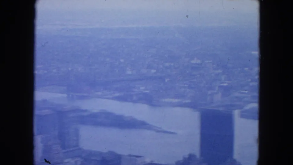 Blue-tinted New York aerial home-movie frame looking toward waterfront, shoreline, and distant urban buildings in haze.