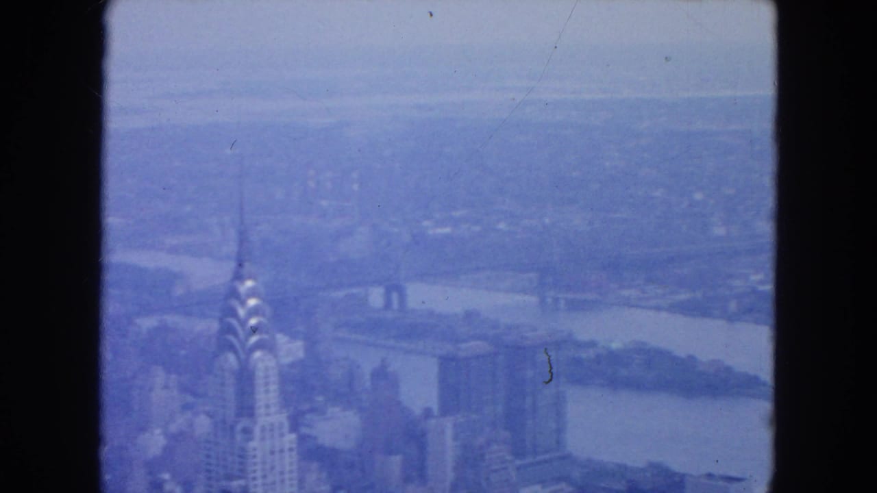 1954 New York City home-movie frame showing the Chrysler Building, distant skyline, and East River in a blue-tinted aerial view.