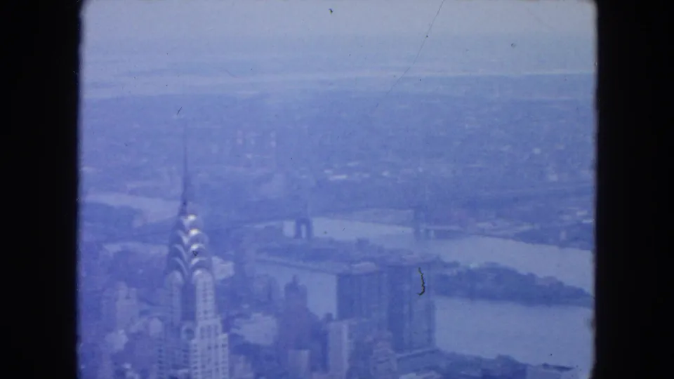 1954 New York City home-movie frame showing the Chrysler Building, distant skyline, and East River in a blue-tinted aerial view.