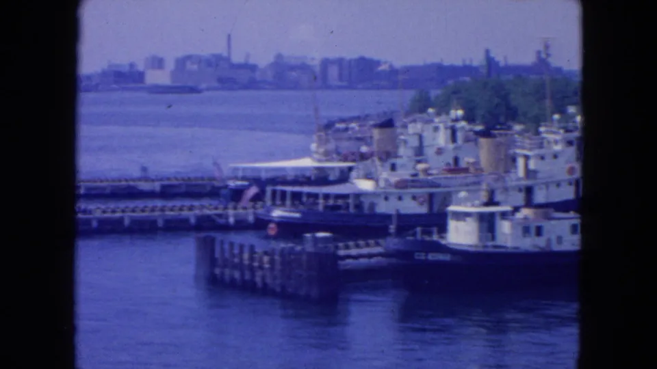 1954 New York City waterfront view with tugboats, pier structures, open river, and distant shoreline in blue-tinted 8mm footage.