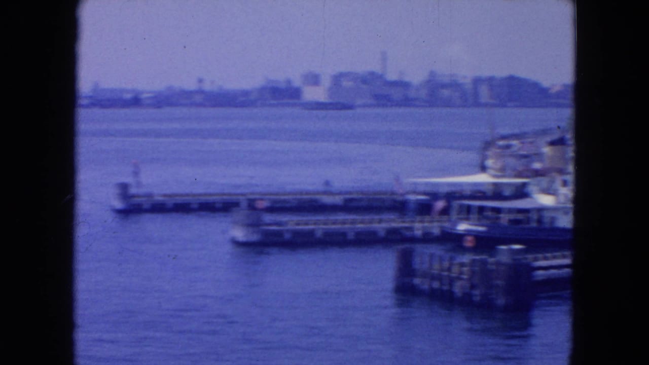 1954 8mm view across New York harbor, showing piers in the foreground and a soft distant skyline under a hazy sky.