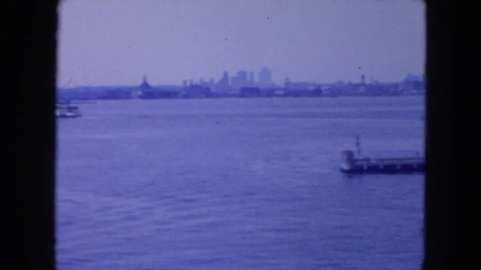 Wide 1954 New York harbor view with calm water, distant skyline, and a small pier visible near the right frame edge.