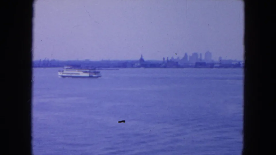 A ferry moves across New York harbor in front of the distant 1954 skyline, seen through soft blue 8mm color.