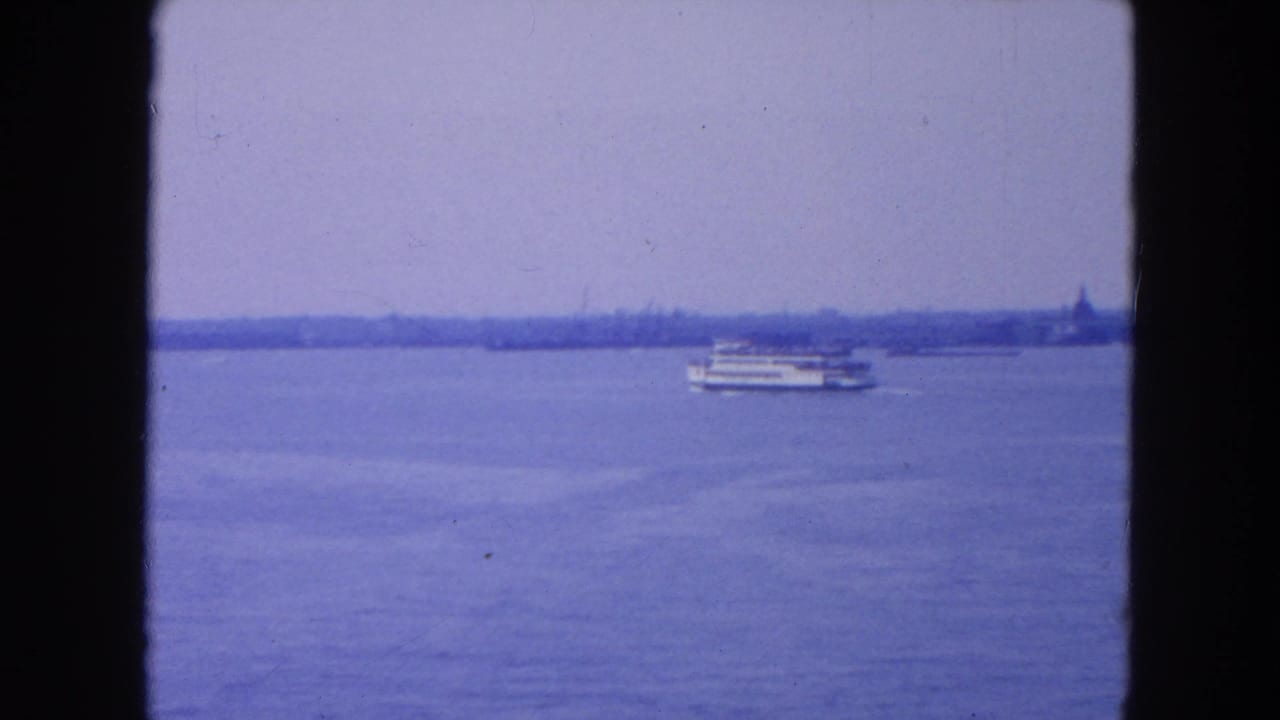 1954 8mm New York City harbor view with blue water, a distant passenger boat, and a hazy shoreline framed by film gate edges.