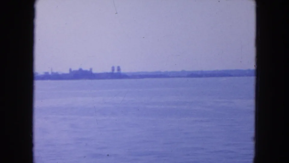 1954 8mm view from the water toward a hazy New York City shoreline with low buildings and open harbor in front.