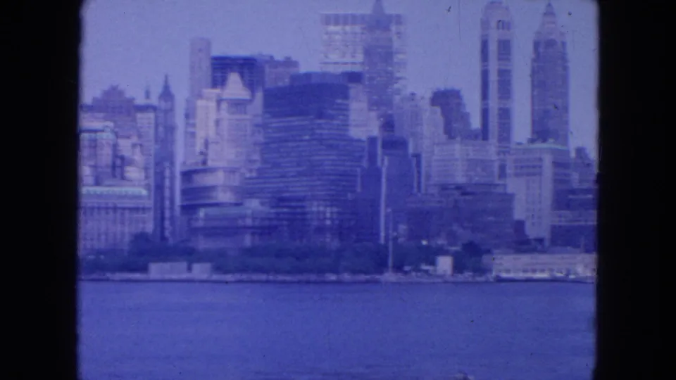1954 8mm view of the New York City skyline across water, with tall mid-century buildings under a hazy blue sky.
