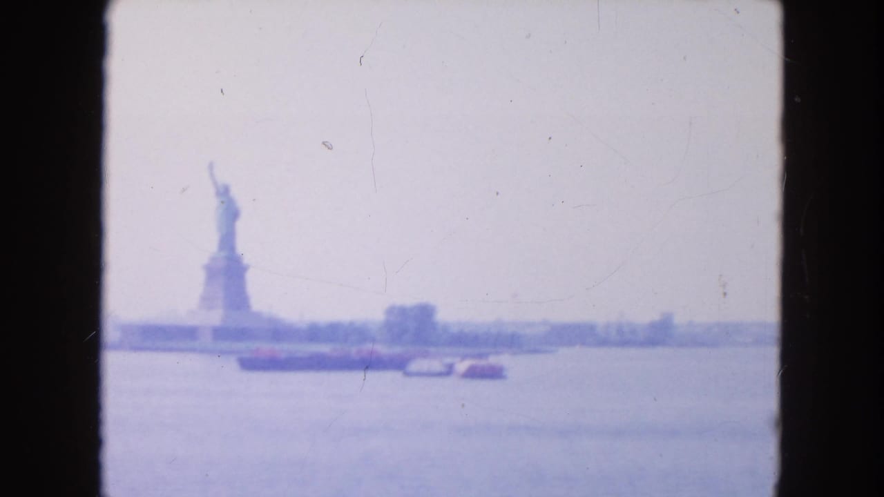 1954 8mm view of the Statue of Liberty seen across New York Harbor, with a low barge and pale sky in soft focus.