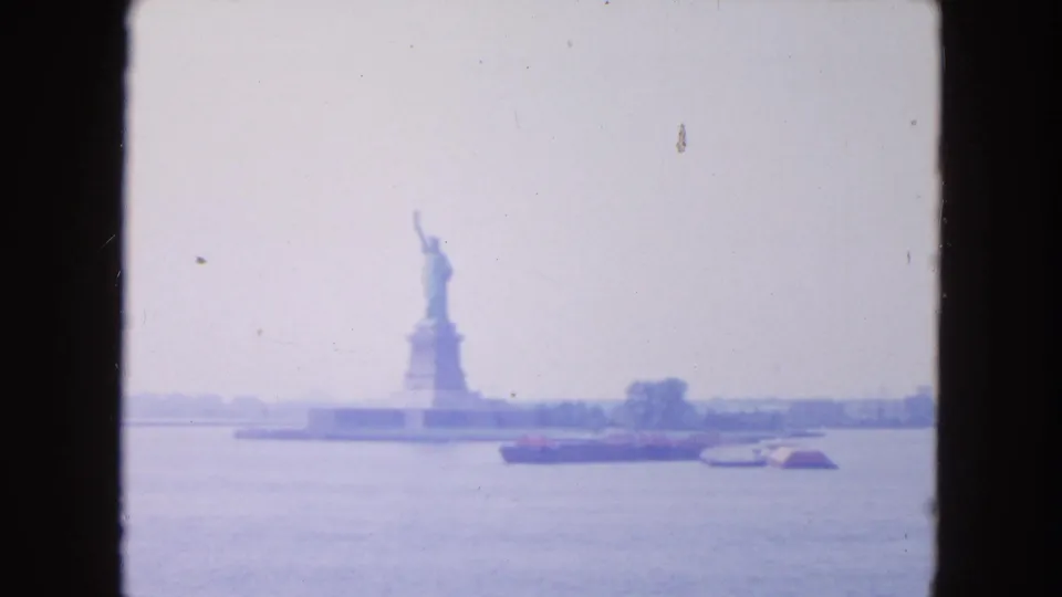 1954 home-movie frame with the Statue of Liberty in the distance, harbor water in front, and small craft near the shoreline.