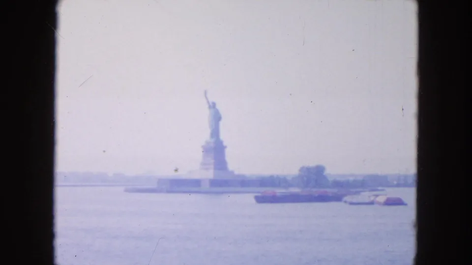 Faded 1954 8mm view of the Statue of Liberty from across New York Harbor, with boats and shoreline trees below a pale sky.