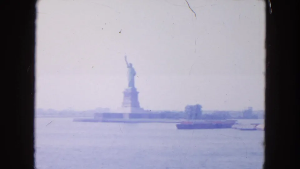 1954 home-movie frame showing the Statue of Liberty from the water, with a low barge and faded blue-purple 8mm tones.