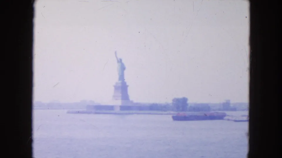Final 1954 8mm frame of the Statue of Liberty across New York Harbor, with water, shoreline, and a red barge visible.