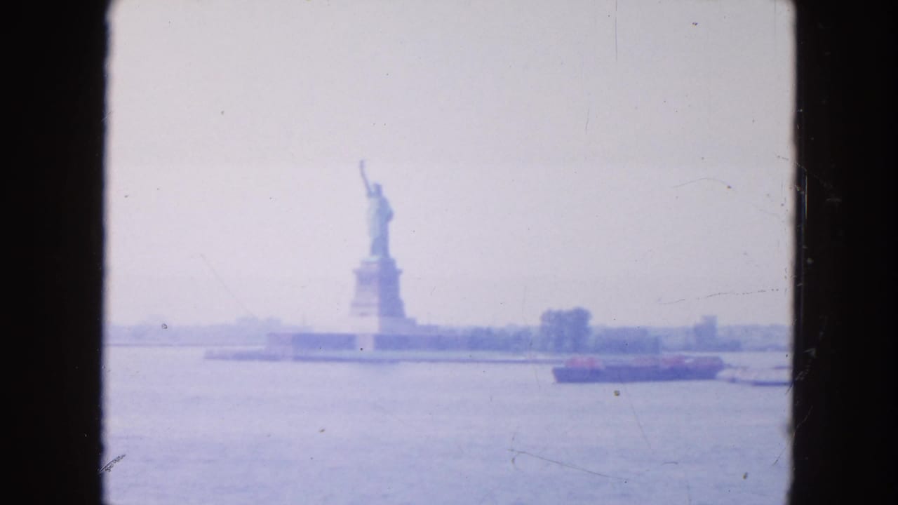 1954 New York Harbor view of the Statue of Liberty from the water, with a low cargo ship crossing the foreground in soft haze.