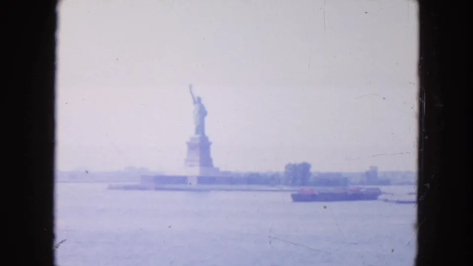 1954 New York Harbor view of the Statue of Liberty from the water, with a low cargo ship crossing the foreground in soft haze.