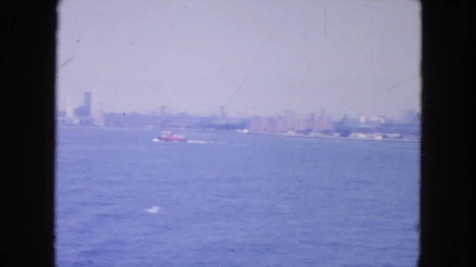 New York Harbor view from the water, with a small red boat moving before a distant shoreline and faint buildings.