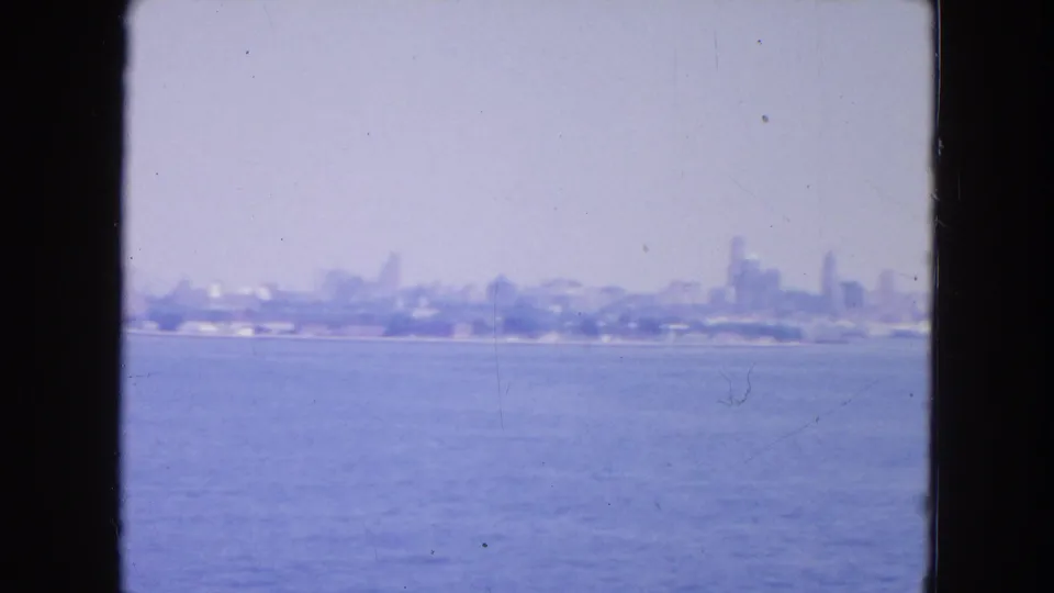 1954 New York City skyline seen hazily across a wide harbor from behind dark window or porthole edges.