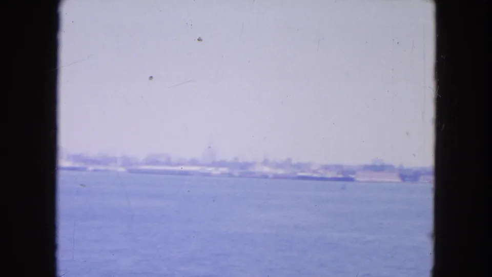 A low New York harbor shoreline sits beyond open water, with the skyline reduced to faint shapes in the haze.