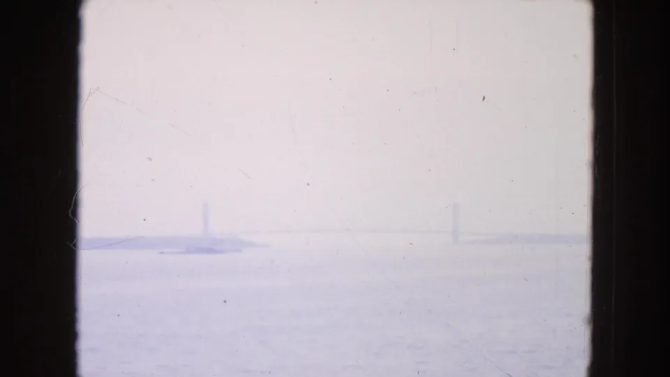 Soft 8mm view across New York harbor water toward a distant shoreline and faint bridge-like forms in heavy haze.