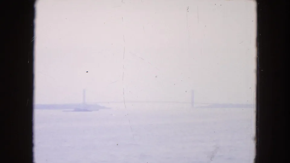 Washed, bluish 8mm frame of harbor water and a distant shoreline with faint vertical structures barely visible through haze.