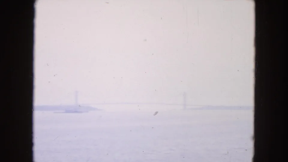 1954 New York City 8mm frame showing a pale, hazy harbor view with distant barges and indistinct bridge-like verticals.