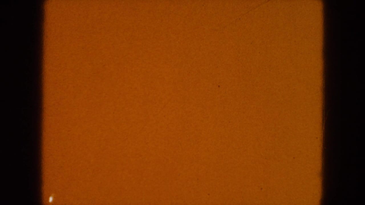 1954 8mm source frame showing warm amber exposure, black film edges, surface speckles, and a small light mark near the lower left.