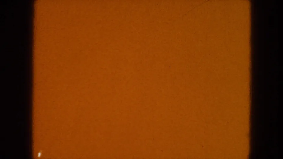 1954 8mm source frame showing warm amber exposure, black film edges, surface speckles, and a small light mark near the lower left.