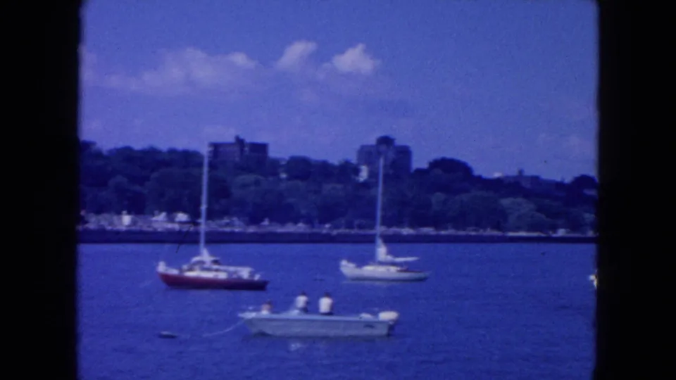 1954 New York City 8mm frame showing sailboats and a low motorboat on the river, with trees and distant buildings along the shore.