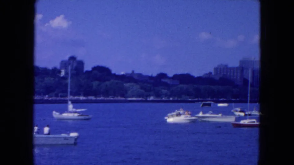 1954 New York City waterfront frame with small recreational boats on the river and a wooded urban shoreline in the background.