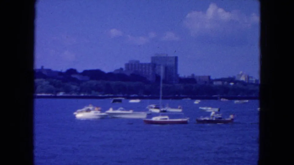 1954 New York City 8mm frame showing a cluster of small boats on the river with trees and taller buildings in the background.