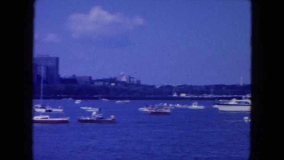Wide 1954 New York City river view with multiple small boats spread across the water and a distant urban shoreline under a blue sky.