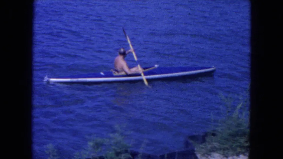 A shirtless adult man paddles a blue kayak across New York City harbor water in this 1954 8mm home-movie frame.
