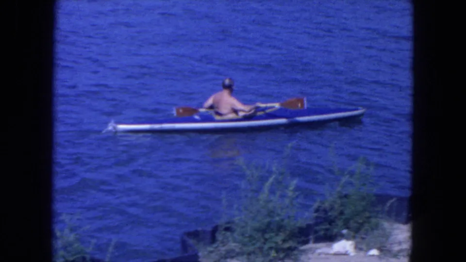 A shirtless adult kayaker is seen from behind on blue harbor water, with distant boats and shoreline visible in 1954 New York City footage.