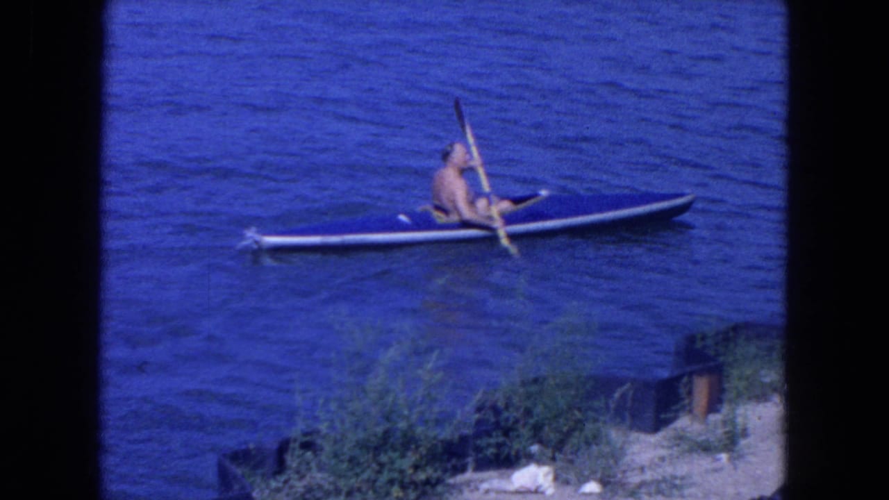 1954 New York City home-movie frame showing a shirtless man paddling a blue kayak on calm water near shoreline greenery.
