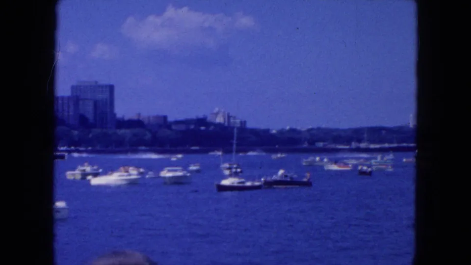 1954 New York City harbor view with small boats on blue water, sailboat masts, and distant buildings along the waterfront.