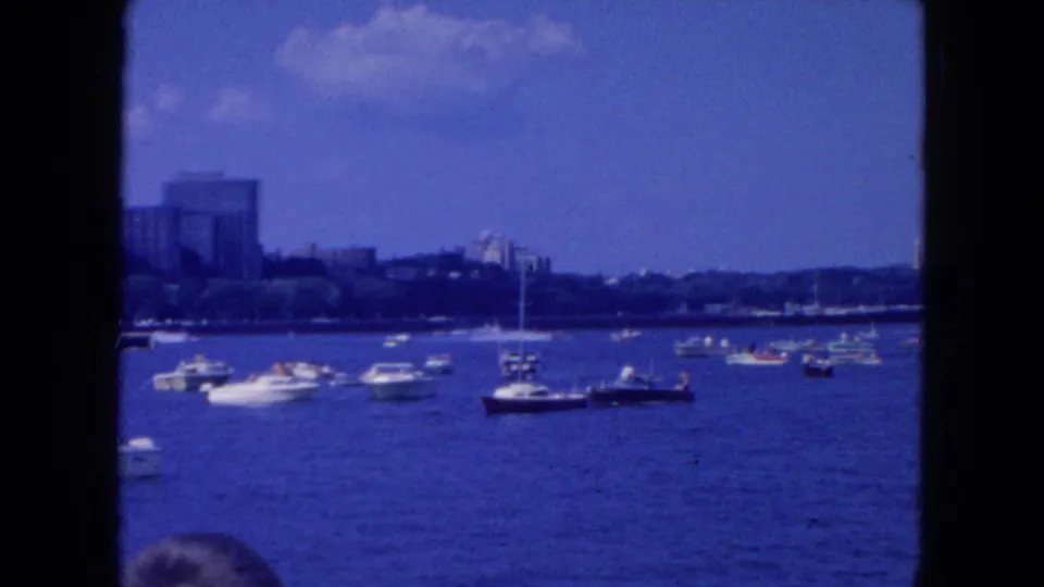1954 New York City home-movie frame showing a wide harbor view with motorboats, sailboats, distant buildings, and a partial head at the lower edge.
