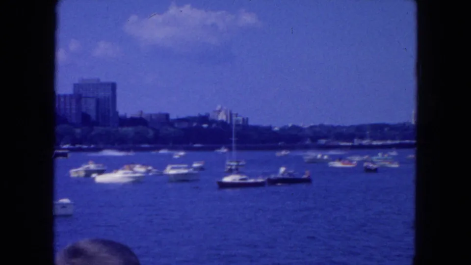 1954 New York City harbor scene with boats spread across blue water, sailboat masts, distant waterfront buildings, and a partial head at the bottom edge.