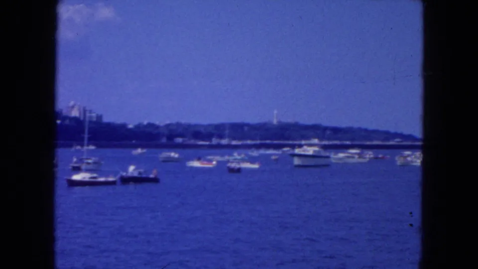 1954 New York City waterfront frame showing scattered boats, a low distant shoreline, and blue-tinted 8mm film texture.