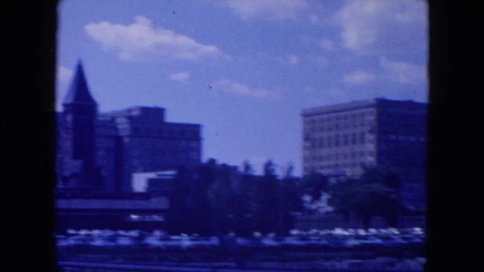 Soft 1954 New York City view of waterfront buildings, including a dark towered structure, trees, and open sky.