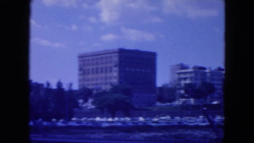 1954 New York City home-movie frame with a large rectangular building, trees, and waterfront edge blurred by movement.