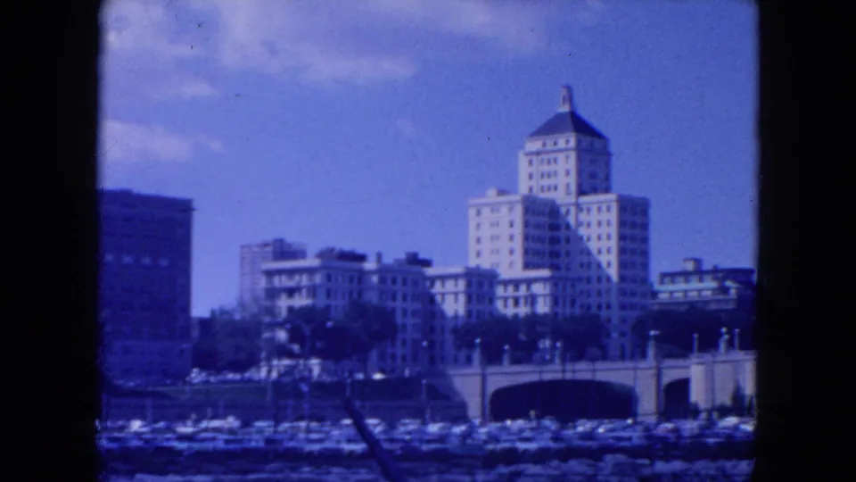 1954 New York City view of multi-story buildings near bridge arches, with waterfront activity low in frame.