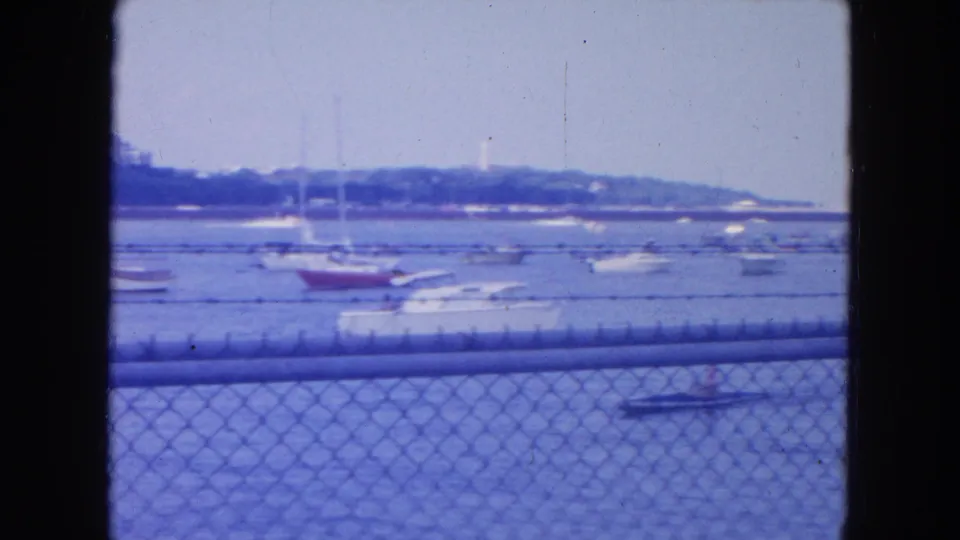1954 New York City waterfront scene with boats on blue water, chain-link fence in front, and a hazy shoreline beyond.