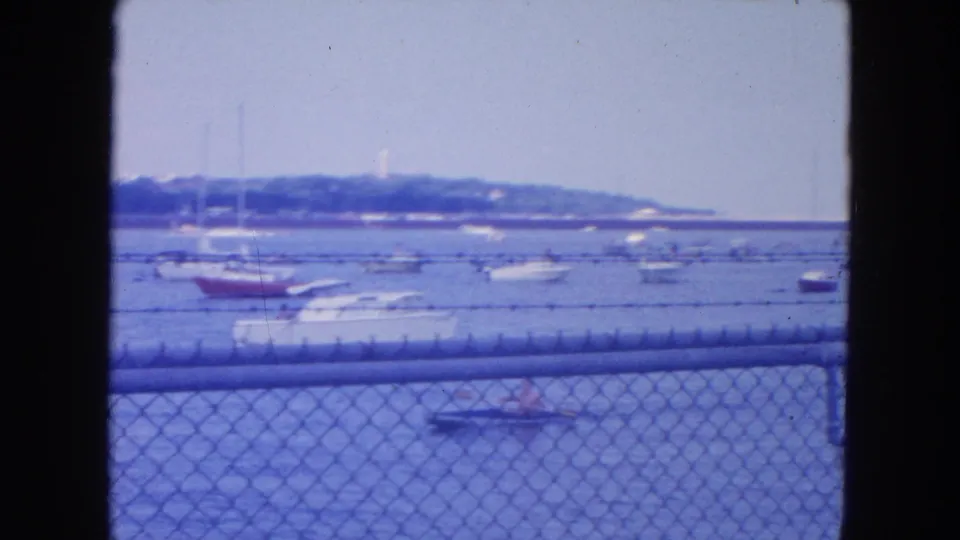 1954 New York City home-movie frame showing moored small boats and sailboats beyond a chain-link waterfront fence.